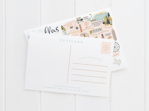 A blank postcard for address and stamp rests atop an Onderkast Studio Dallas Map Postcard, which features iconic city landmarks, all displayed on a white wooden surface.