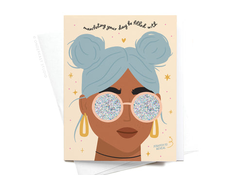 The Manifesting Good Vibes Scratch-Off Greeting Card by Onderkast Studio features an illustration of a person with light blue hair in two buns, holographic glitter sunglasses, gold earrings, and a star-and-heart background.