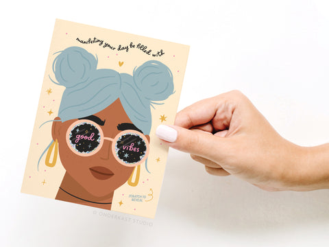 A hand holds the Manifesting Good Vibes Scratch-Off Greeting Card by Onderkast Studio, featuring a blue-haired woman with good vibes sunglasses, stars, holographic glitter, and manifesting your day is filled with at the top.