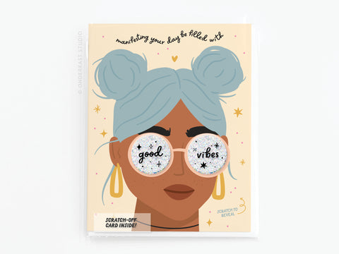 Onderkast Studios Manifesting Good Vibes Scratch-Off Greeting Card features a blue-haired person with buns, gold earrings, and sunglasses with scratch-off “good vibes” lenses. Text above: “Manifesting your day be filled with.”.