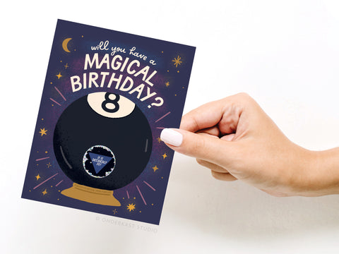 A hand holds the Onderkast Studio Magic 8-Ball Birthday Scratch-Off Greeting Card, featuring a Magic 8-Ball and “Will you have a magical birthday?” surrounded by stars and sparkles on a dark background.