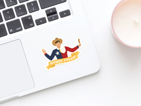 A laptop with the Onderkast Studio Howdy Folks Sticker—featuring a smiling cowboy in a Texas flag shirt holding a Corny Dog beneath a “Howdy Folks!” banner—next to a pink candle on a white surface.