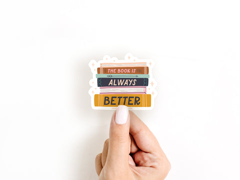 A hand with white nail polish holds The Book Is Always Better Sticker by Onderkast Studio, a vinyl sticker shaped like a stack of books, against a plain white background. Proudly printed in the USA.