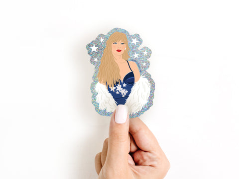 A hand with light nail polish holds the Onderkast Studio TS Midnights Glitter Sticker featuring a blonde woman in a blue outfit with white fur, set against a plain white background—a fun must-have for Swifties.
