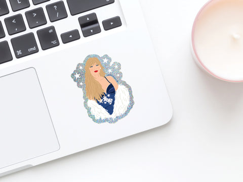 A laptop keyboard featuring the Onderkast Studio TS Midnights Glitter Sticker, showing a woman with long blonde hair in a blue dress and white fur, beside a pink candle on a white surface—perfect for any Swiftie’s setup.