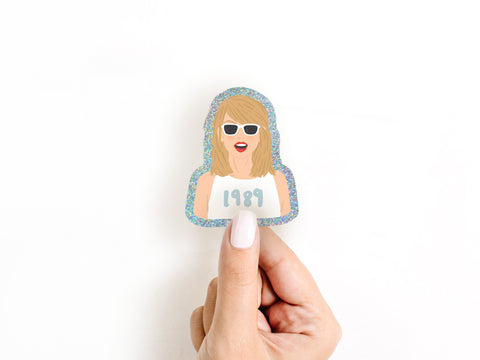 A hand with white nail polish holds the Onderkast Studio TS 1989 Glitter Sticker, a weatherproof vinyl sticker featuring a woman in 1989 shirt, blonde hair, red lipstick, and white sunglasses, shown against a white background.