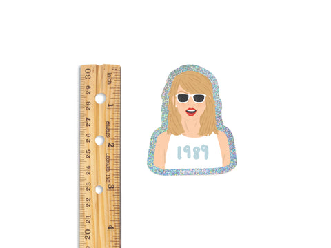 The Onderkast Studio TS 1989 Glitter Sticker, about 3 inches tall and made from weatherproof glitter vinyl, features a woman with blonde hair, white sunglasses, and a white top. It is shown next to a ruler for scale.