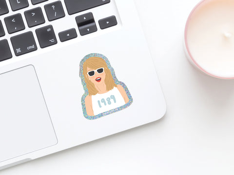 The TS 1989 Glitter Sticker by Onderkast Studio, featuring a woman in white sunglasses, red lipstick, and a 1989 shirt, decorates this laptop keyboard next to a pink candle.