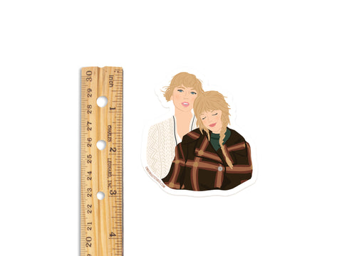 The Onderkast Studio TS Folkevermore Sticker features two women—one in a white sweater, the other in a brown plaid jacket—leaning together. This weatherproof vinyl sticker is about 3 inches tall.