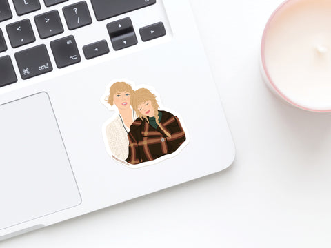 A laptop displays the TS Folkevermore Sticker by Onderkast Studio—depicting two blonde women in cozy outfits. Nearby, a white candle in a pink holder sits on the white desk.