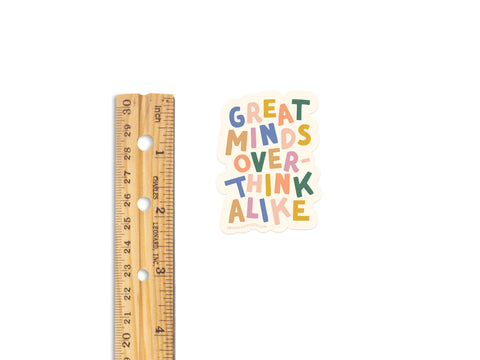 A wooden ruler next to an Onderkast Studio Great Minds Overthink Alike Sticker—printed in the USA on weatherproof vinyl with playful, multicolored letters on a white background.