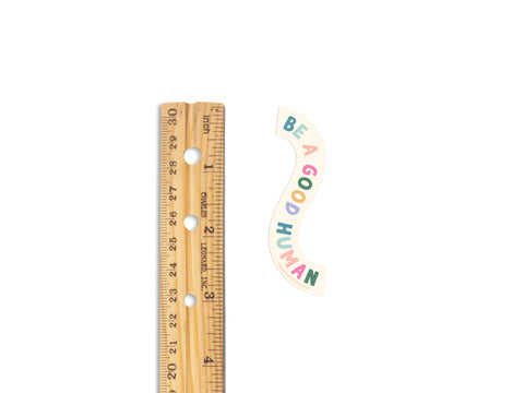 A wooden ruler sits beside the Onderkast Studio Be A Good Human Sticker, featuring colorful letters on white vinyl. This weatherproof sticker is perfect for spreading kindness wherever you place it.