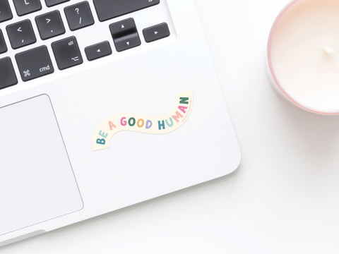 A Be A Good Human Sticker by Onderkast Studio, featuring colorful letters, brightens any workspace. This weatherproof vinyl sticker is perfect for adding positivity and charm to your laptop or desk.