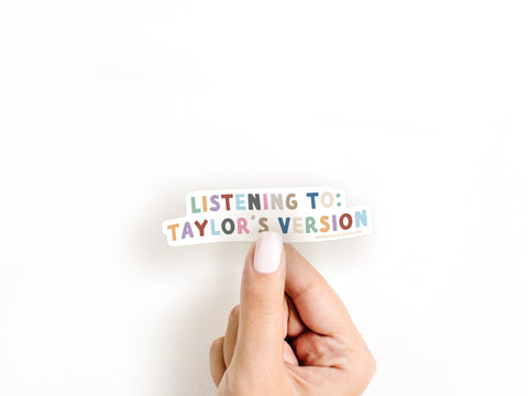 A hand with light nail polish holds the Onderkast Studio Listening To Taylor’s Version Sticker, featuring bold, colorful letters and celebrating Taylor’s re-recordings, against a plain white background.