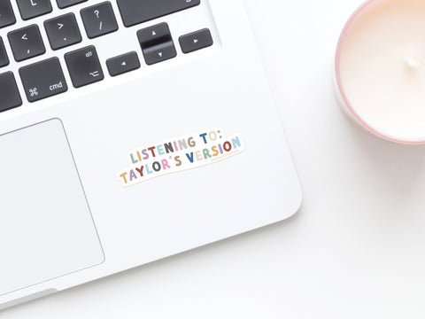 A laptop with an Onderkast Studio Listening To Taylor’s Version Sticker in colorful letters near the keyboard. Close by, a white candle in a pink holder—ideal for fans of Taylors re-recordings.