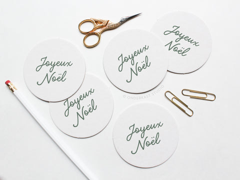 The Joyeux Noël Coaster Set by Onderkast Studio features five round letterpress coasters in green script on durable pulp board, arranged on a white surface with a pencil, gold scissors, and paperclips.