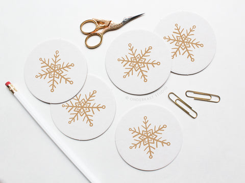 The Snowflake Coaster Set by Onderkast Studio features five round white coasters with gold snowflake designs, paired with gold scissors, paper clips, and a white pencil—beautifully arranged to showcase their handmade quality.