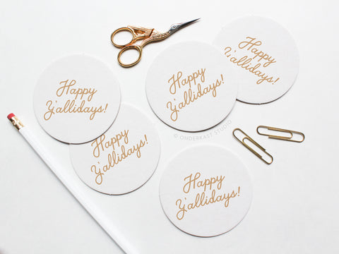 The Happy Y’allidays Coaster Set by Onderkast Studio features five round white letterpress coasters in gold script, styled with a white pencil, gold scissors, and paperclips on a white surface to showcase their handmade quality.