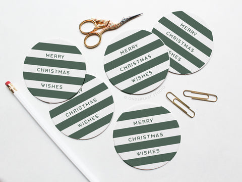 The Merry Christmas Wishes Coaster Set by Onderkast Studio features round pulp board coasters with green and white stripes and “Merry Christmas Wishes” text, displayed on a white surface with a pencil, scissors, and gold paper clips.