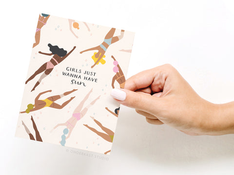 A pale hand with manicured nails holds the Onderkast Studio Girls Just Wanna Have Sun Greeting Card – FINAL STOCK, featuring vibrant illustrations of women swimming and the phrase GIRLS JUST WANNA HAVE SUN.