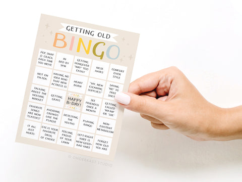 A hand holds the Onderkast Studio Getting Old Bingo Greeting Card, featuring a playful 5x5 grid with funny aging signs on a clean white background.