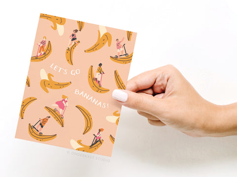 A hand holds the Lets Go Bananas Greeting Card – FINAL STOCK by Onderkast Studio, featuring lively people on scooters and bananas—a premium card for vibrant celebrations, set against a peach background.