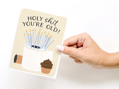 A hand holds the Onderkast Studio “Holy Shit You’re Old!” greeting card, featuring a toilet paper roll with candles and a poop emoji cupcake, perfect for milestone birthdays.
