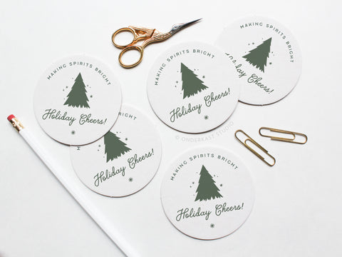 The Holiday Cheers Coaster Set by Onderkast Studio features five round letterpress coasters with a Christmas tree design and “Making Spirits Bright, Holiday Cheers!” text, arranged on a white surface with office supplies to highlight handmade quality.