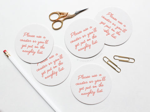 The Naughty List Coaster Set by Onderkast Studio features four round white letterpress coasters with “Please use a coaster or you’ll get put on the naughty list” in red script, made from durable pulp board.