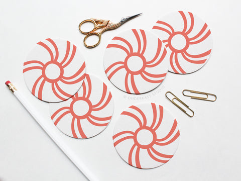 The Peppermint Candy Coaster Set by Onderkast Studio features round white letterpress coasters with red swirls, shown on a white surface beside a white pencil, gold paperclips, and ornate gold-handled scissors to highlight their handmade quality.