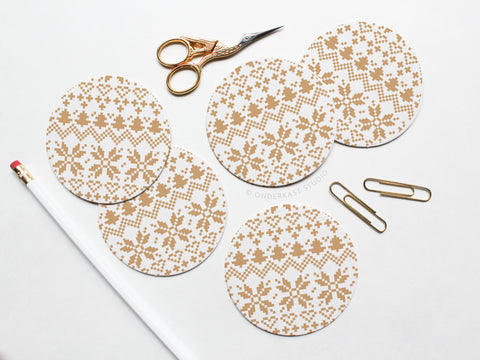 The Holiday Cross Stitch Coaster Set by Onderkast Studio features five round pulp board coasters with a beige snowflake pattern, shown on a white surface with gold scissors, two gold paperclips, and a white pencil with red eraser.