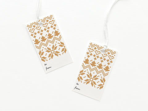 Set of two white Holiday Cross Stitch Gift Tags by Onderkast Studio, made from card stock with gold Nordic snowflake patterns, to: and from: in handwritten-style font at the bottom, and attached white strings.