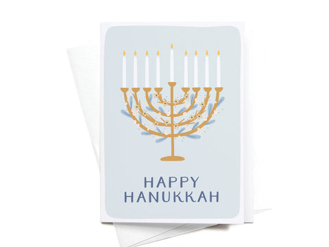 The Onderkast Studio Happy Hanukkah Floral Menorah Folded Greeting Note Set of 10 features a golden menorah with all candles lit and sparkles on a light blue background, with “HAPPY HANUKKAH” below, printed in the USA.