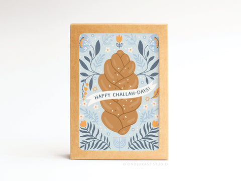 The Happy Challah-days! Folded Greeting Note Set of 10 by Onderkast Studio features an illustration of braided challah bread with a “Happy Challah-Days!” banner, floral and menorah designs, on a light blue background. Printed in the USA.