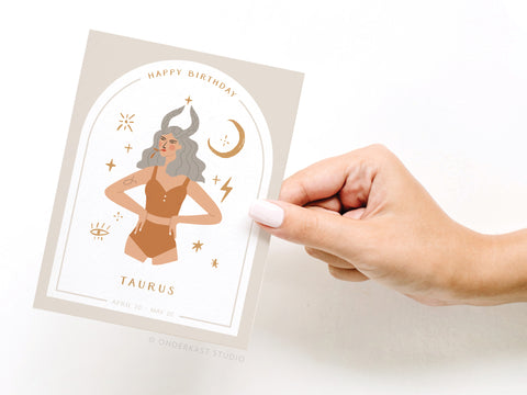 A hand holds the Happy Birthday Taurus Zodiac Greeting Card by Onderkast Studio, featuring an illustrated woman with bull horns, stars, and celestial symbols. The card reads Happy Birthday Taurus, April 20 - May 20.
