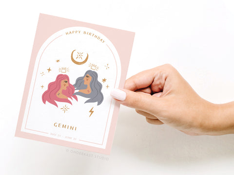 A hand holds the Onderkast Studio Happy Birthday Gemini Zodiac Greeting Card in pastel pink, featuring two illustrated women, stars, a moon, Happy Birthday, and May 21-June 20 dates at the bottom.
