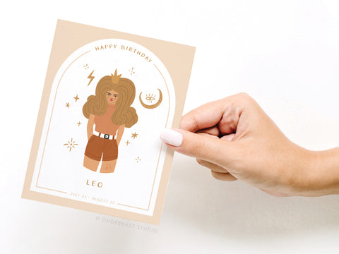 A hand with painted nails holds the Onderkast Studio Happy Birthday Leo Zodiac Greeting Card, showing a cartoon woman with big hair, stars, zodiac symbols, and July 23–August 22 dates on a beige background—ideal for Leos.