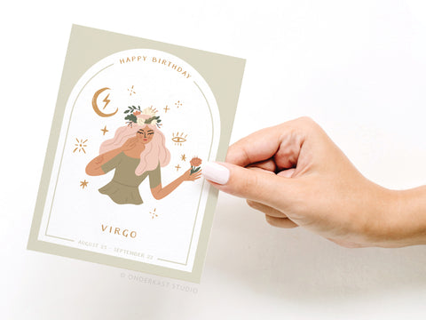 A hand with light nail polish holds the Happy Birthday Virgo Zodiac Greeting Card by Onderkast Studio, featuring an illustrated woman with flowers, astrological symbols, and Virgo dates (August 23 - September 22).