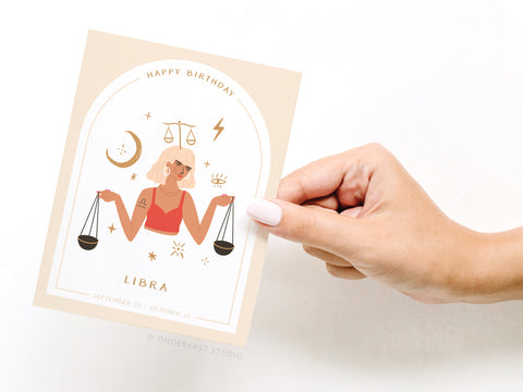 A hand holds the Onderkast Studio Happy Birthday Libra Zodiac Greeting Card, featuring an illustrated woman with short hair, earrings, sunglasses, Libra scales, stars, and a crescent moon. Printed in the USA. Text: “Happy Birthday Libra” and “September 23 - October 22.”.