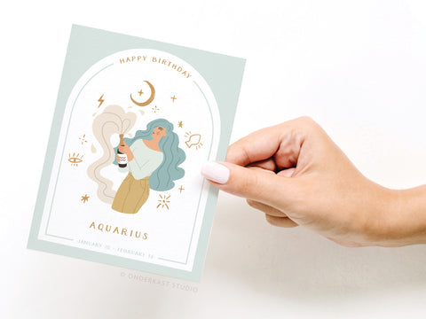A hand holds the Onderkast Studio Happy Birthday Aquarius Zodiac Greeting Card, featuring a woman with teal hair holding a bottle among stars and zodiac symbols. The card reads “Happy Birthday Aquarius” and lists the dates January 20 - February 16.