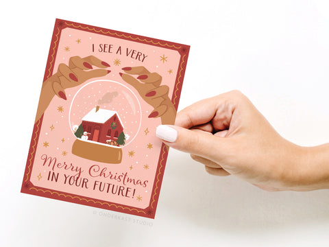 A hand holds Onderkast Studios I See a Very Merry Christmas In Your Future card, featuring a snow globe with a red house, stars, and mystical hands. The festive greeting appears on this premium holiday card.