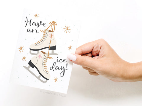 A hand holds the Onderkast Studio Have an Ice Day! Ice Skates greeting card—FINAL STOCK—which features illustrated ice skates with the message Have an ice day! surrounded by stars and dots.