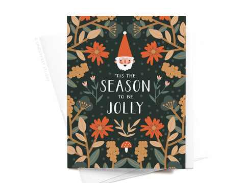 The ’Tis the Season to Be Jolly Greeting Card by Onderkast Studio features a smiling gnome in a red hat with red and yellow flowers on a dark background and the text “Tis the season to be jolly.” Printed in the USA.