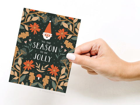 A light-skinned hand with white nail polish holds the Onderkast Studio ’Tis the Season to Be Jolly Greeting Card, featuring floral designs, a Santa face, and festive text on a dark background. Printed in the USA.
