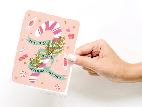 A hand with white nail polish holds the Onderkast Studio Joyful & Sweet Holiday Wishes Candy Cane Greeting Card, featuring greenery and candies on a pink background, printed on premium card stock.