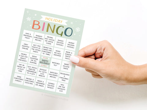 A hand holds the Onderkast Studio Holiday Bingo Greeting Card, featuring a festive title, snowflake decorations, and bingo squares with interactive holiday activities.