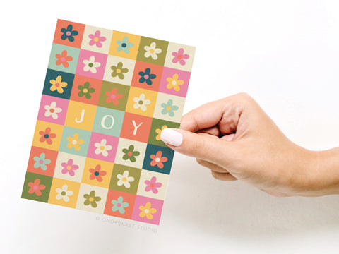 A hand with light nail polish holds the Onderkast Studio Joy Daisies Greeting Card, featuring a grid of daisies and JOY in white on three squares amid the floral pattern.