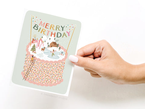 A hand with pale pink nails holds the Onderkast Studio Merry Birthday Cake Greeting Card, featuring a sprinkle cake, winter scene, and festive bunting—perfect for stylish winter birthday celebrations.
