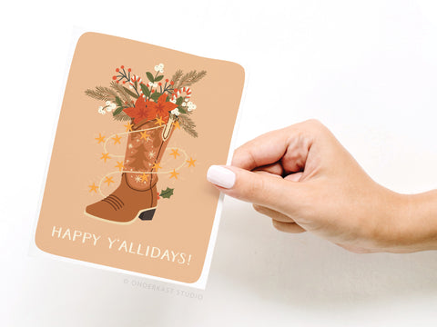 A hand with pale nail polish holds the Onderkast Studio Happy Y’allidays! Cowboy Boot Greeting Card, featuring festive flowers and greenery. “HAPPY Y’ALLIDAYS!” is written in white letters, capturing Southern charm.
