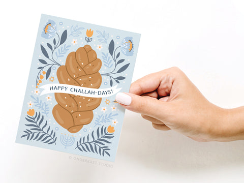 A hand holds Onderkast Studio’s Happy Challah-days! Greeting Card, featuring illustrated challah bread with blue and orange flowers, a menorah at the bottom, and the message “Happy Challah-Days!”.